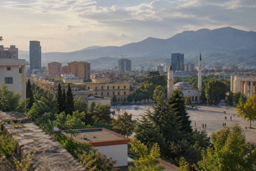 Evening view over the low rooftops and tree canopies of Tirana toward distant mountain ridges