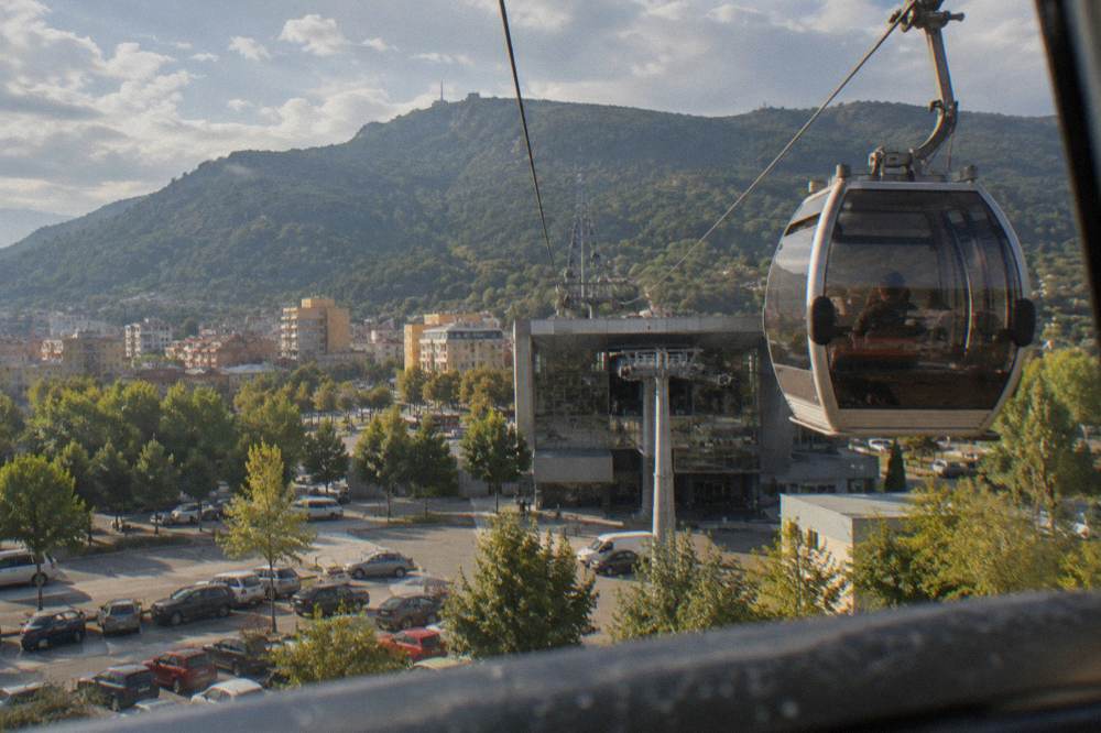 The glass cabin of a cable car rising above Tirana toward the green slopes of Mount Dajti