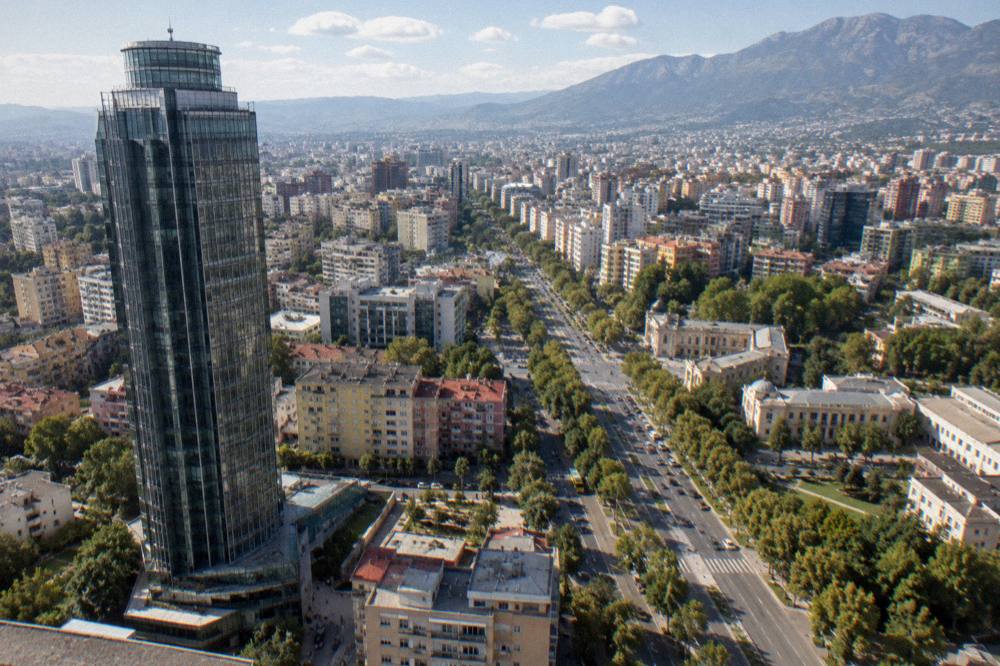 A wide panorama of Tirana's rooftops and boulevards under a clear sky with mountains on the horizon