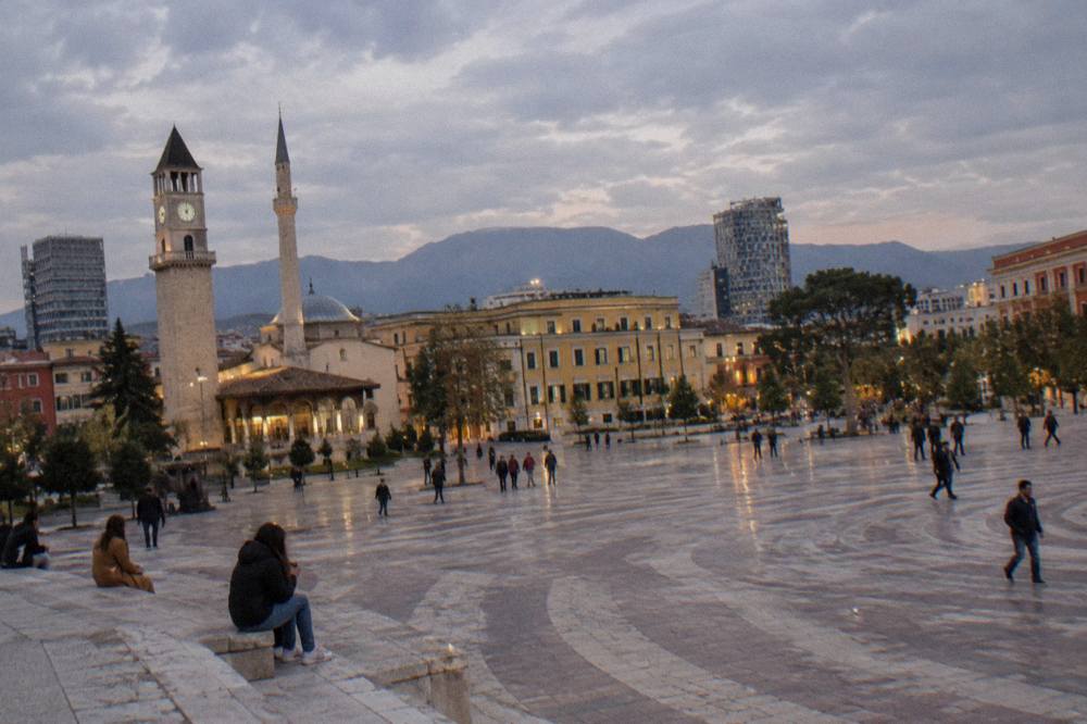 Night view of Tirana with streets and buildings lit up and dark mountain outlines beyond