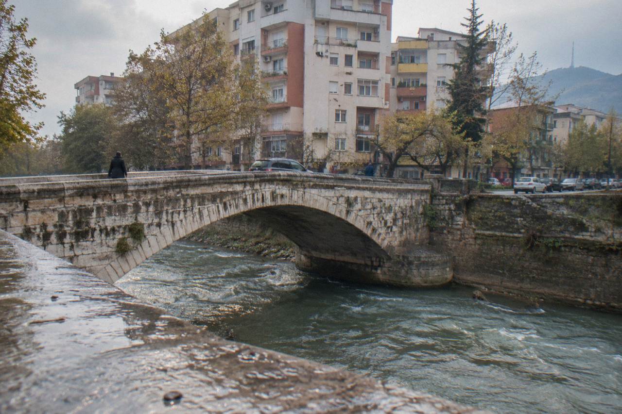 A low stone arch bridge on a quiet street in Tirana with parked cars and apartment buildings behind it