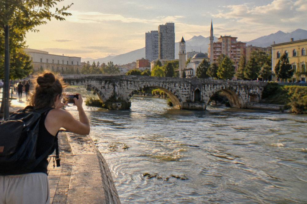 A visitor photographing the stone arch of an old bridge from the waterside path in Tirana