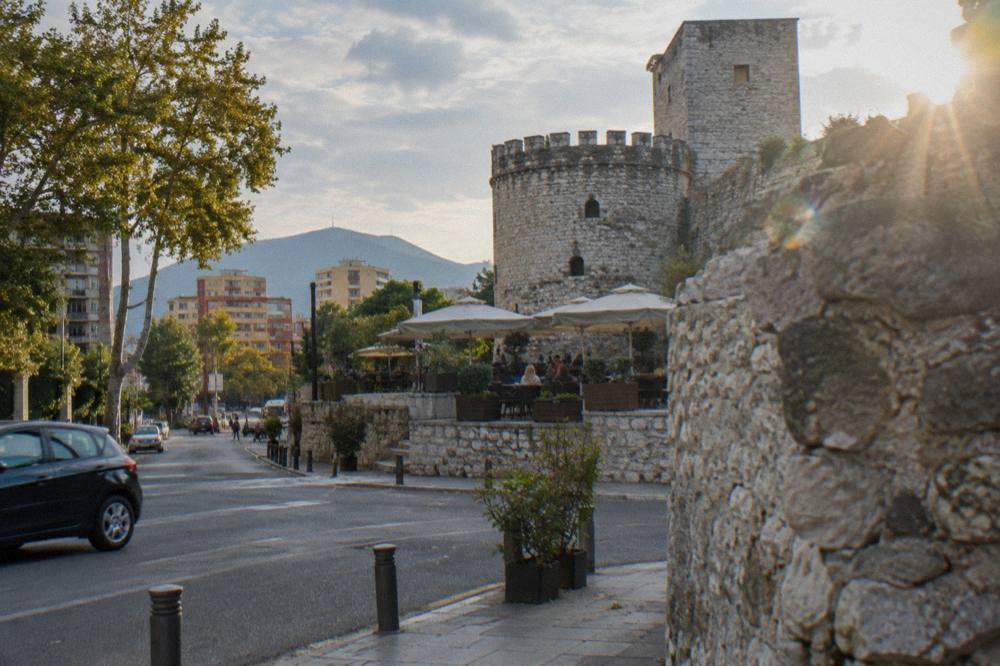 A rough stone wall section and rounded tower of Tirana Castle beside a modern street