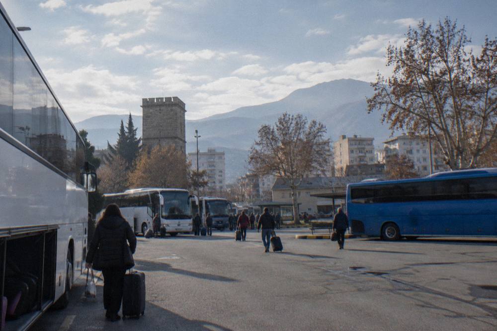 A bus terminal forecourt with intercity buses and passengers with bags