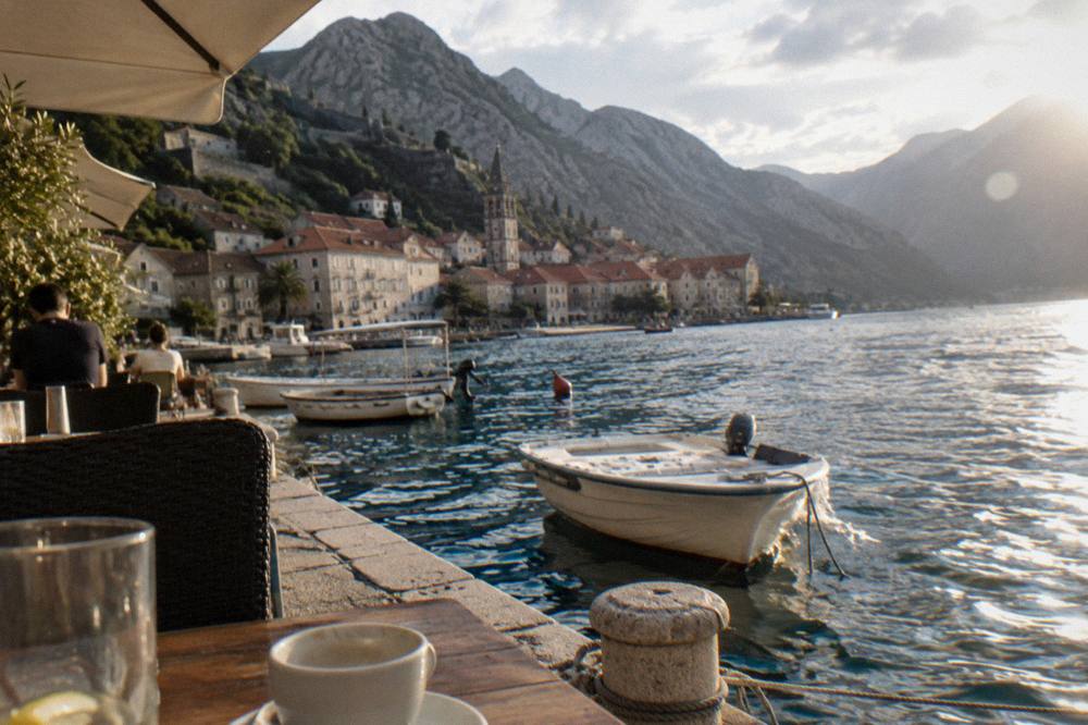Small boats moored along a stone waterfront with cafe tables in the shade