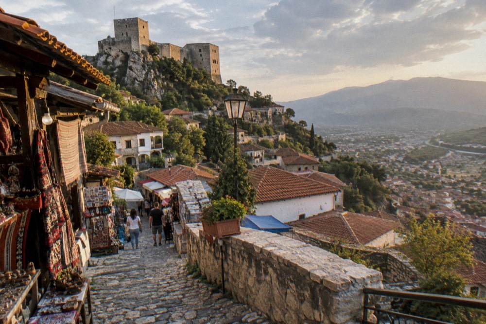 A hill town of old stone houses with orange-tiled roofs climbing a steep slope beneath a fortress on a rocky mountain spur