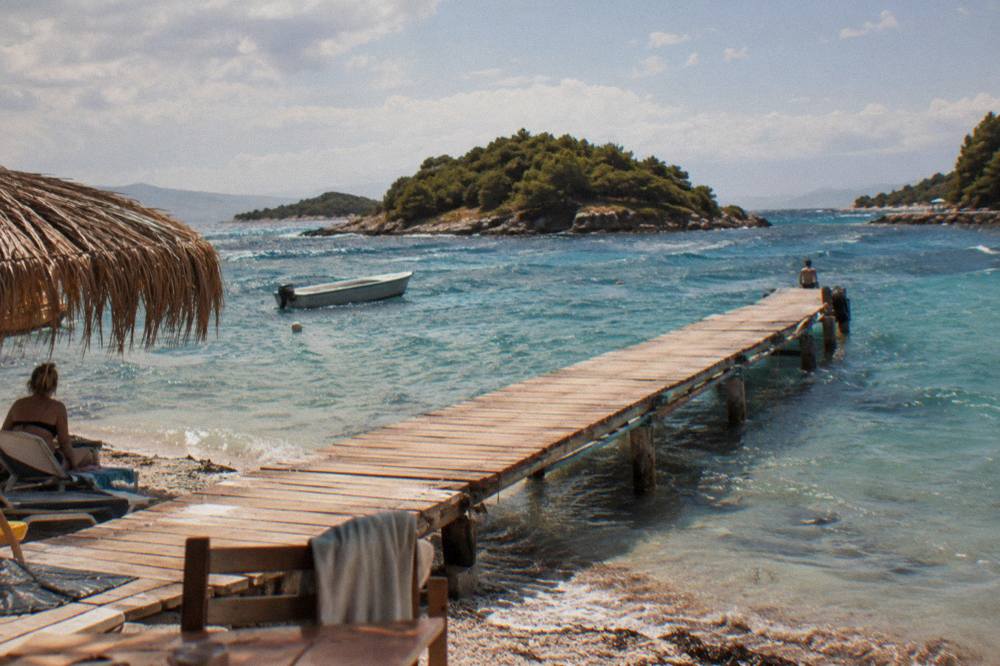 A wooden jetty extending over clear shallow water toward green islands at sunset in Ksamil