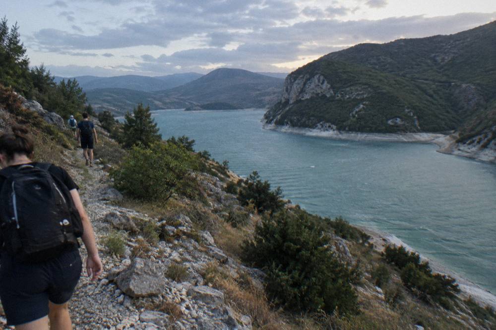 Hikers on a rocky path ascending a dry ridge above deep green water