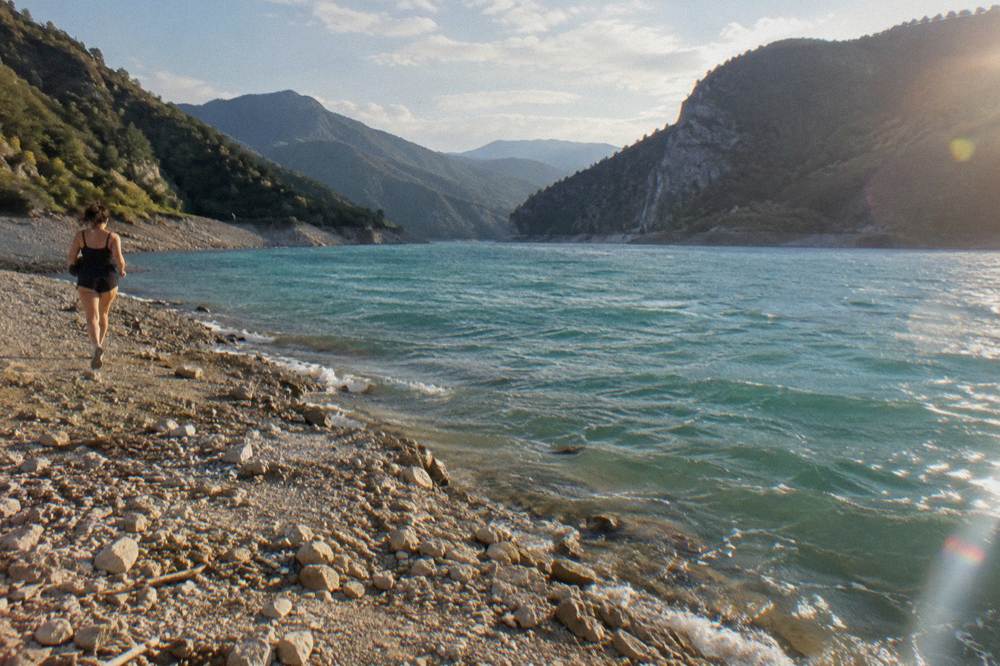 A wide calm reservoir of clear blue-green water ringed by steep wooded hills and bare rock with a quiet shore of pale stones