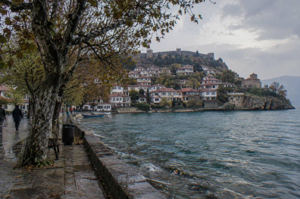 A calm lakeside promenade with plane trees and water reflecting hills