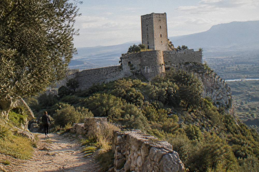 The stone walls and round tower of a hilltop castle seen from the path below