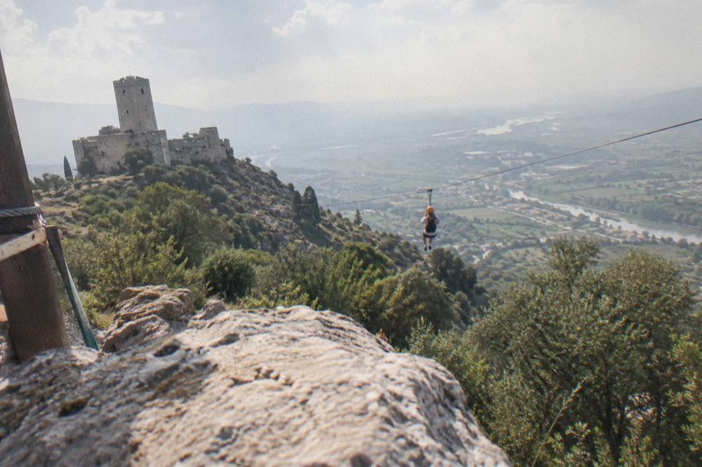A person on a zipline line descending a green hillside toward a valley