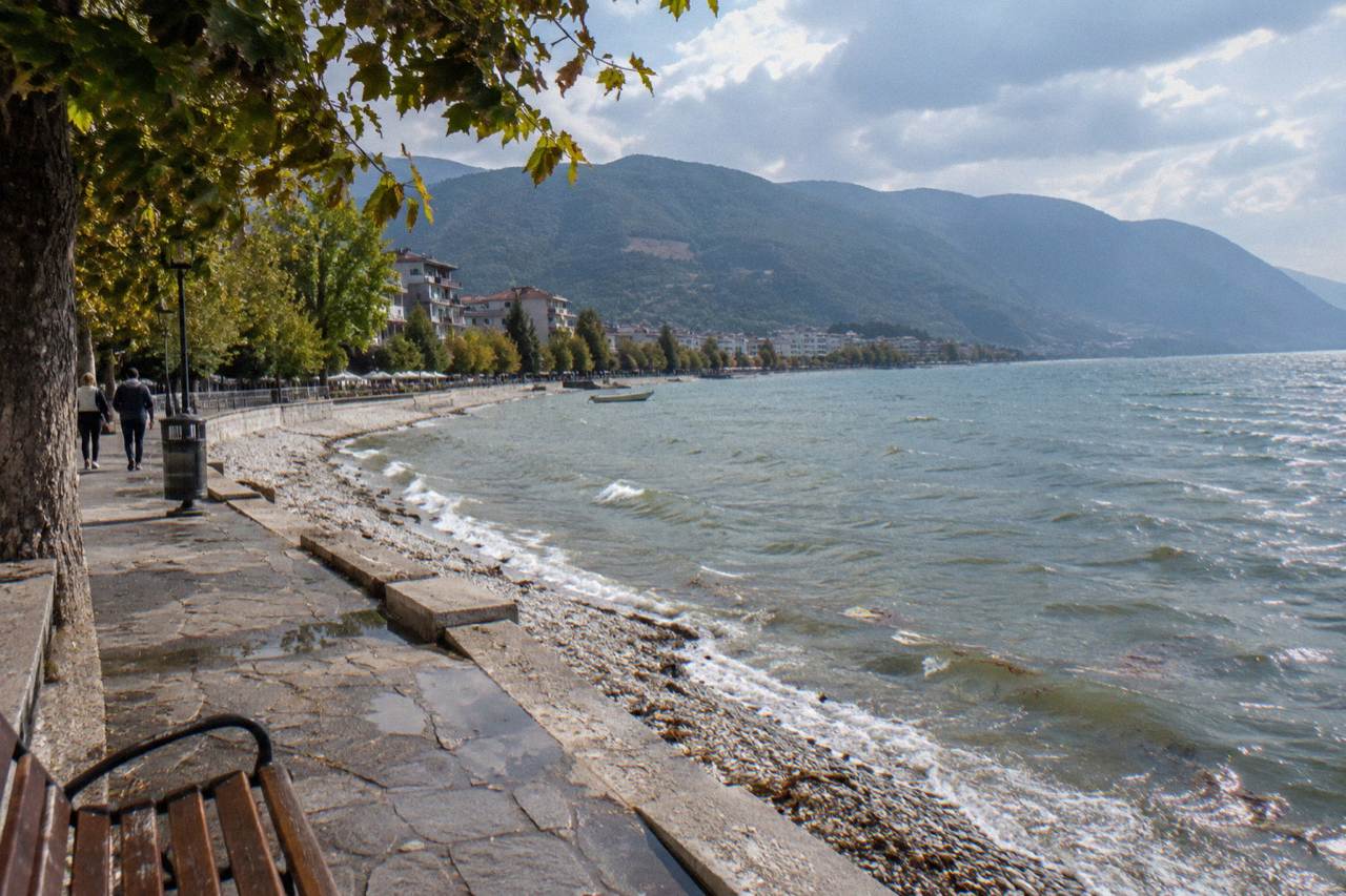 A calm lakeside promenade with low buildings and hills rising across the water
