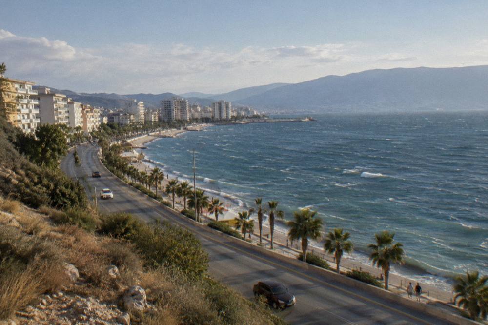 A wide coastal road curving between dry hills and the sea under a clear sky