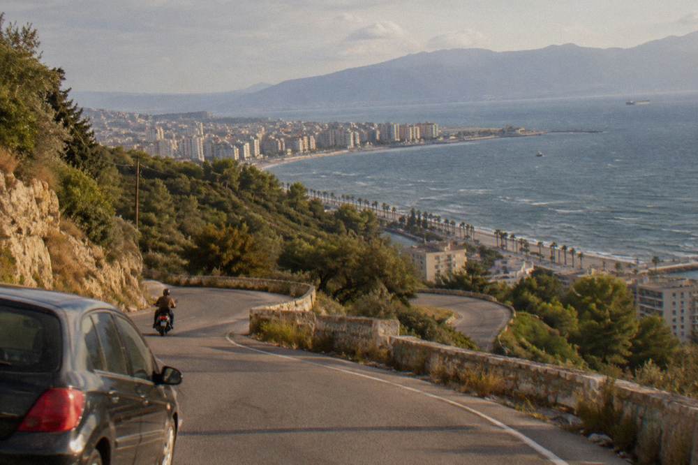 A steep mountain pass road with hairpin bends above forested slopes and distant sea