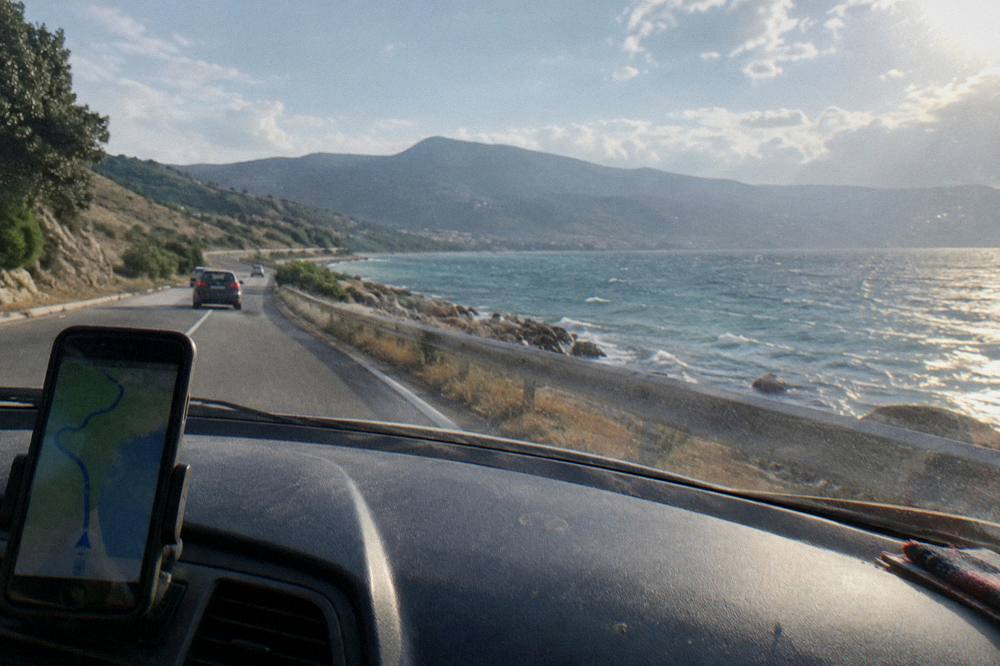 A winding mountain road above a lake with a phone mounted on a car dashboard showing navigation