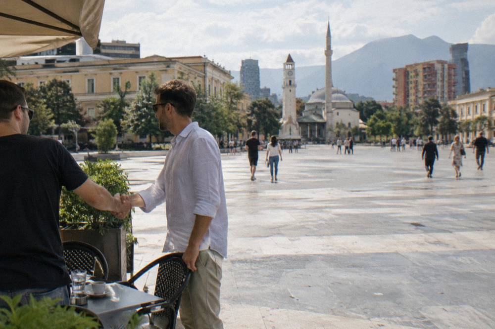 Two friends shaking hands outside a small cafe on a Tirana street