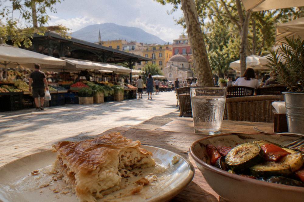 Flaky baked pastry with white cheese and a small bowl of grilled vegetables on a wooden table in soft window light