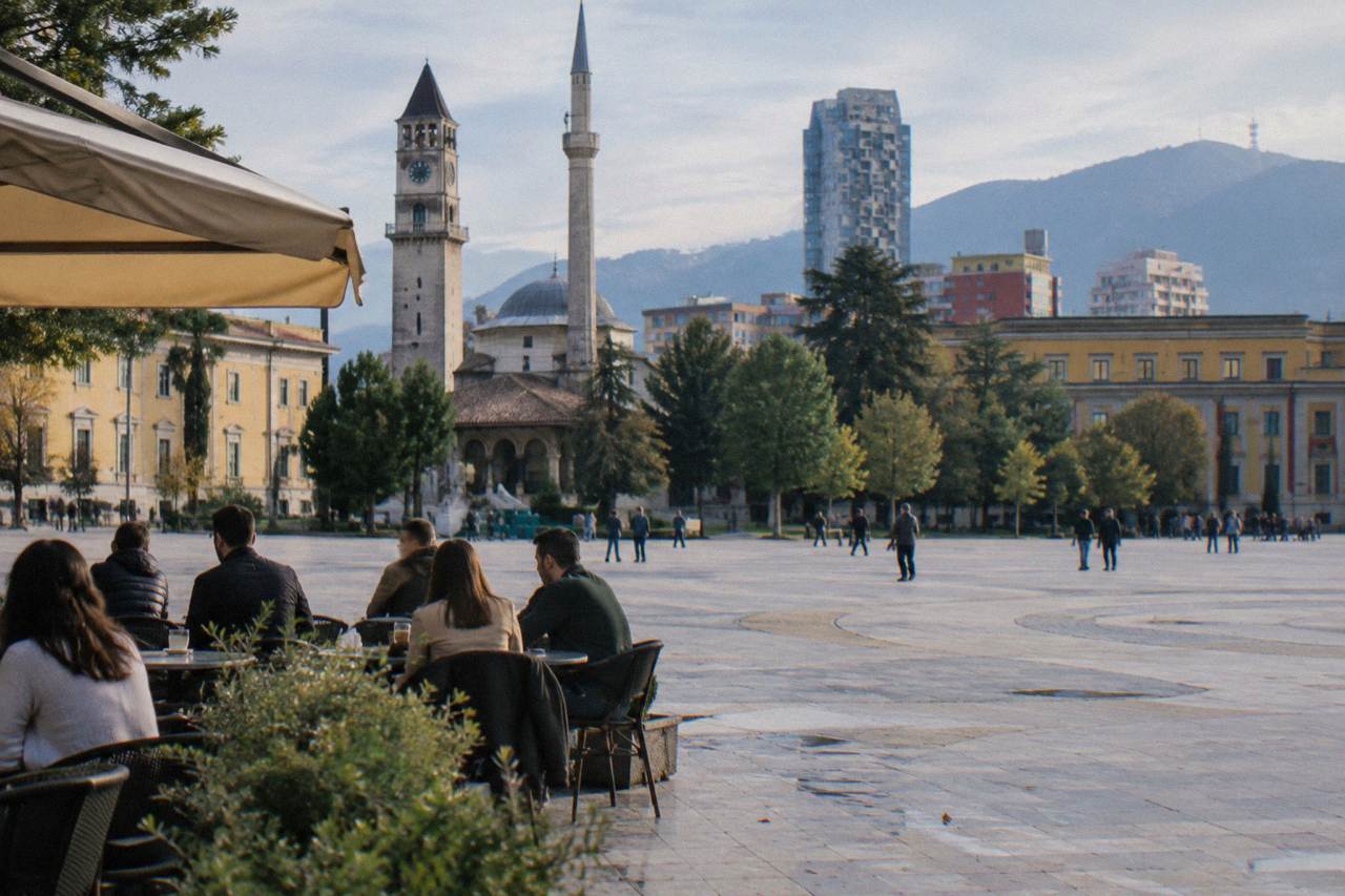 A cafe terrace in central Tirana with people talking at small tables in the morning light