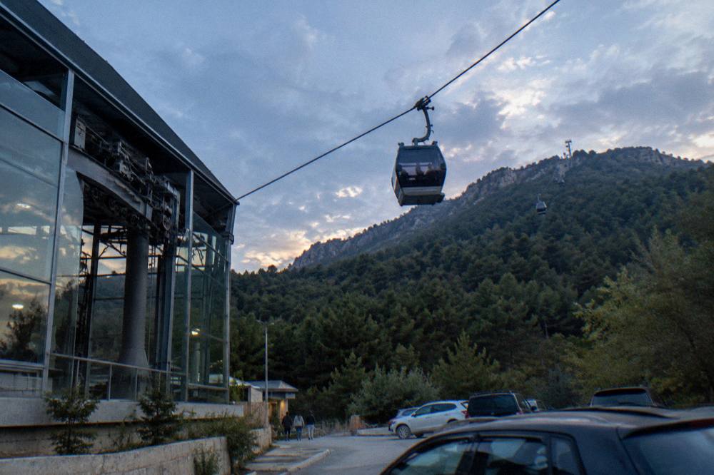 A cable car cabin rising above pine forest toward a mountain ridge