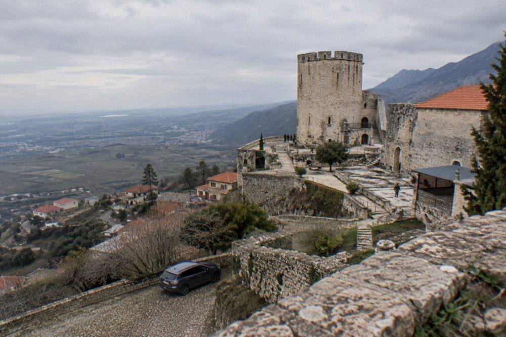 A car parked below the stone walls of Kruja Castle with hills behind