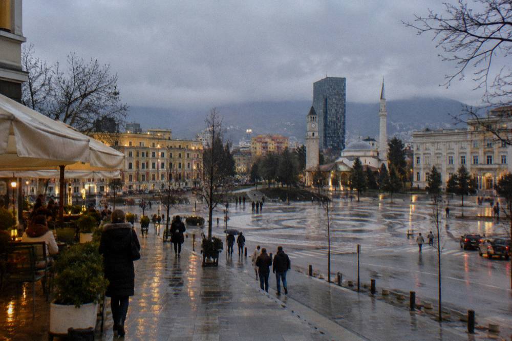 A rainy evening street in Tirana with wet pavement reflecting cafe lights