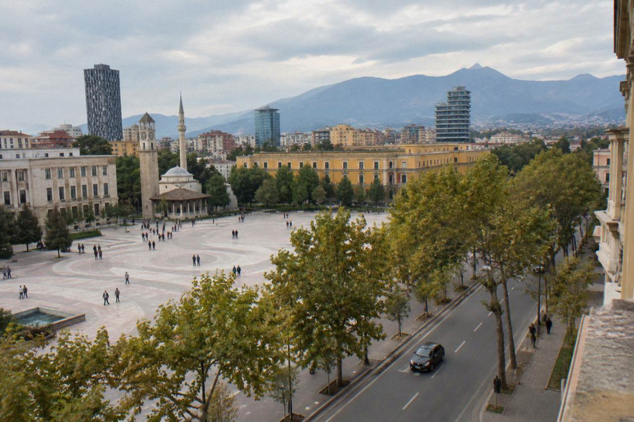 A car driving along a tree-lined boulevard in central Tirana with hills in the distance