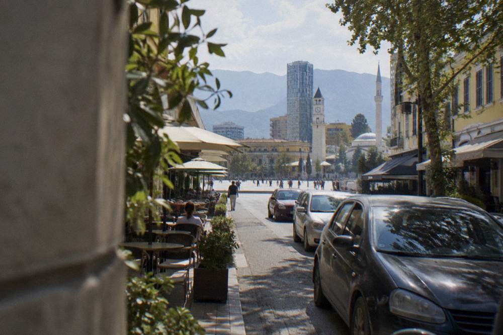 Cars parked along both sides of a narrow street lined with cafes in a Tirana neighbourhood