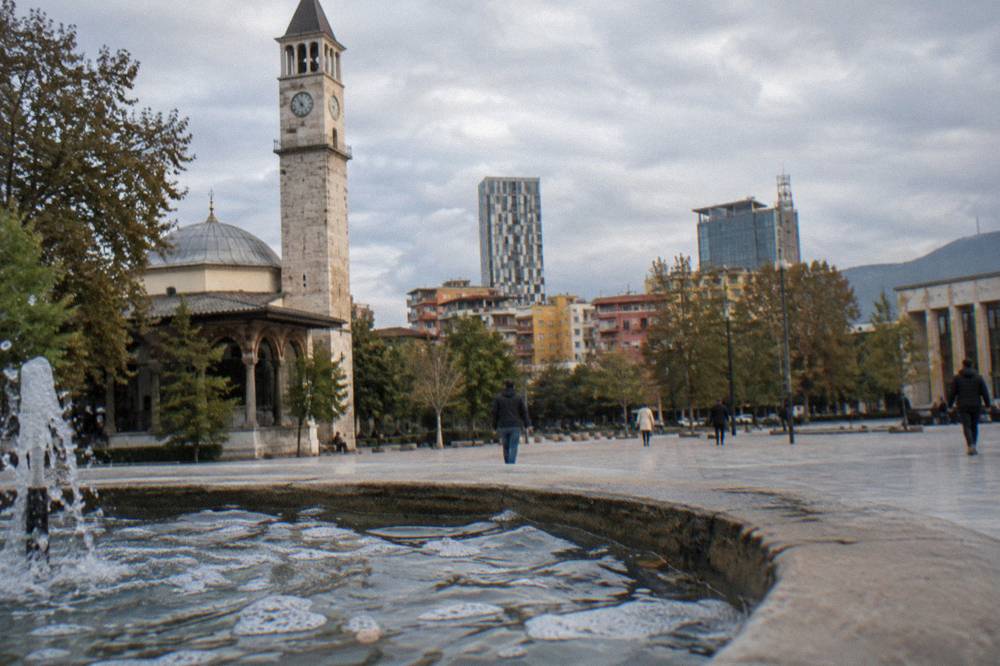A mosque with a clock tower beside it at the edge of a city square in late afternoon light