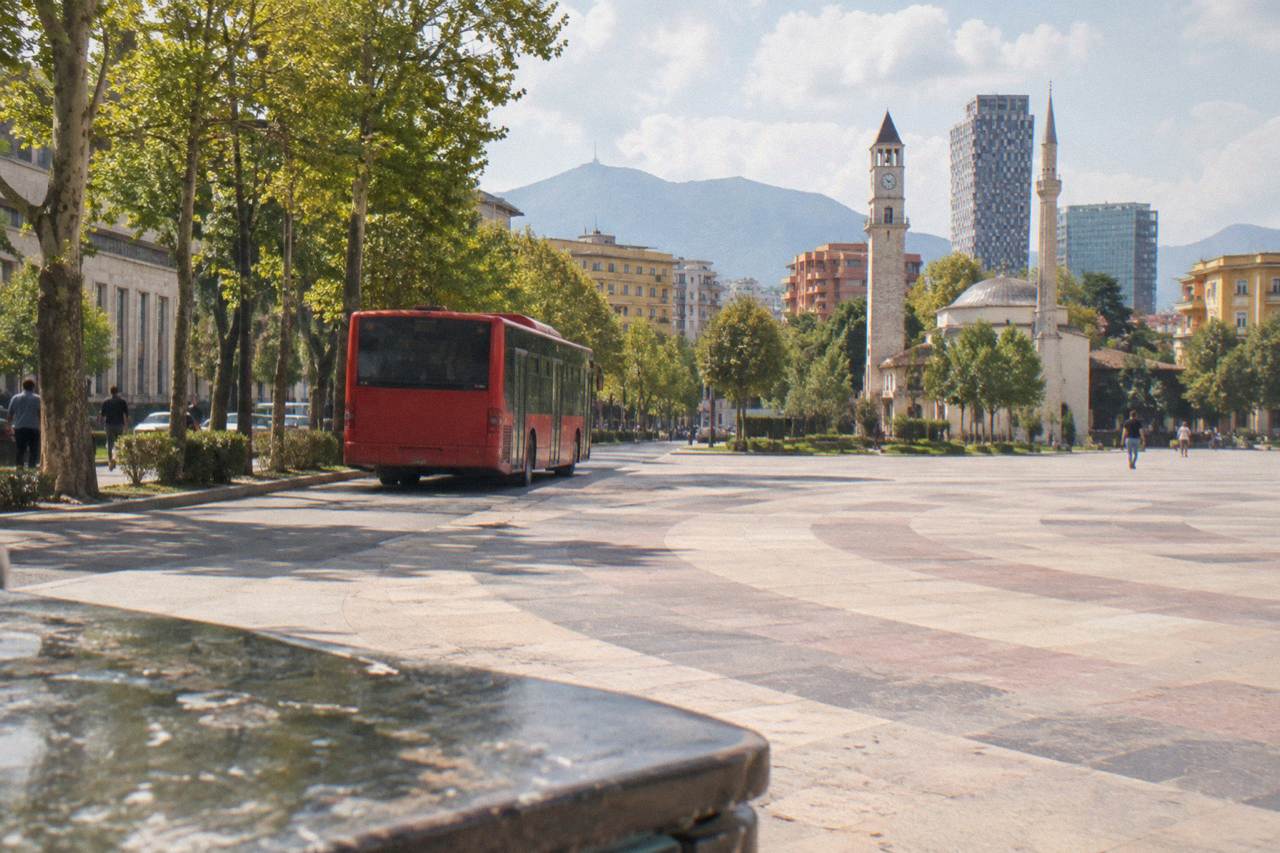 A red Tirana city bus pulling away from a stop on a tree-lined boulevard