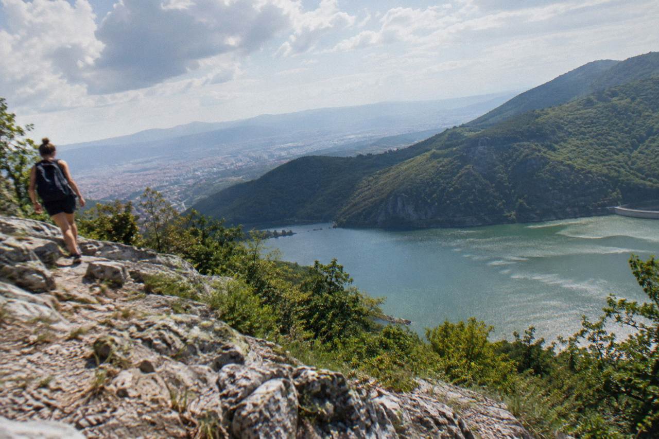 A hiker on a rocky ridge above a green lake reservoir with mountains behind