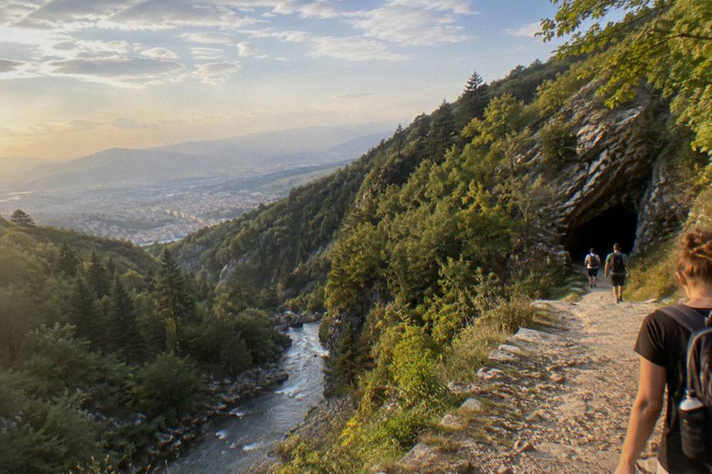 A stone and dirt path along a green river gorge toward a dark cave entrance