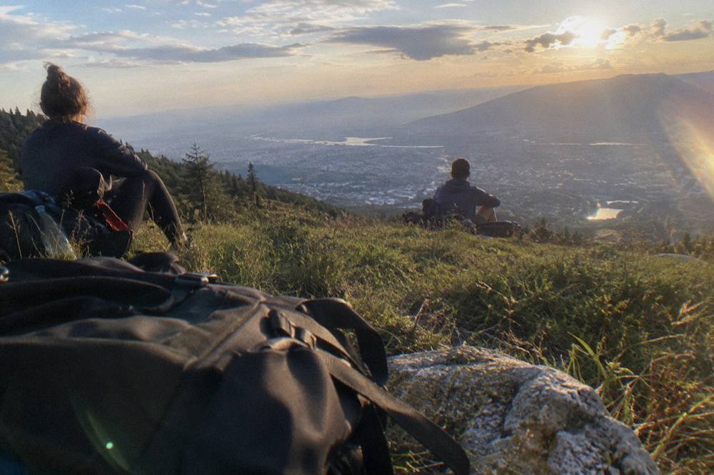 Two hikers resting on a grassy ridge at sunset above a wide valley city