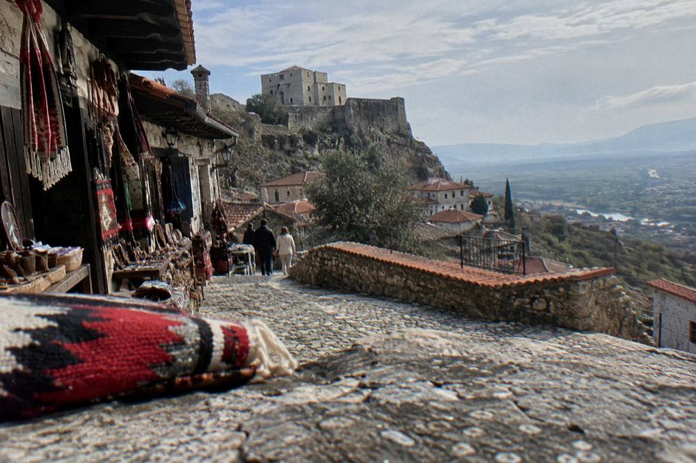 A stone castle wall and red-tiled roofs of Kruja old town above a green valley