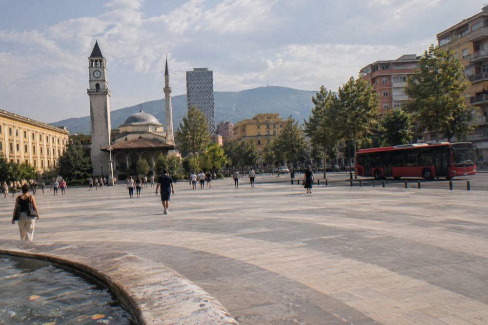 A red city bus on a boulevard in Tirana with apartment blocks and hills behind
