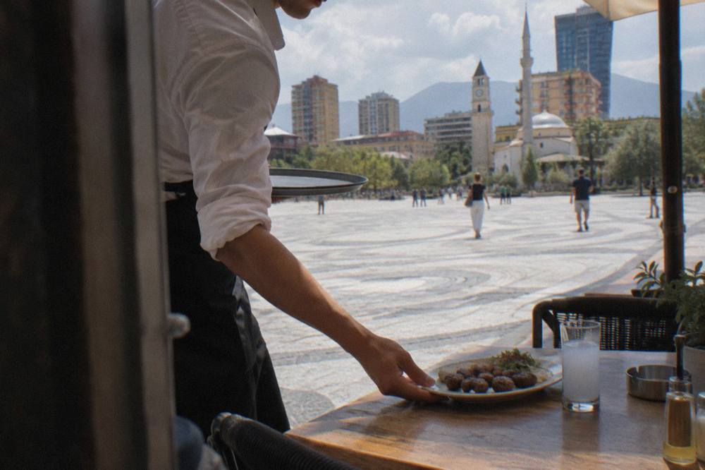 A waiter setting down a plate of grilled meatballs and a glass of raki on a restaurant table in Tirana