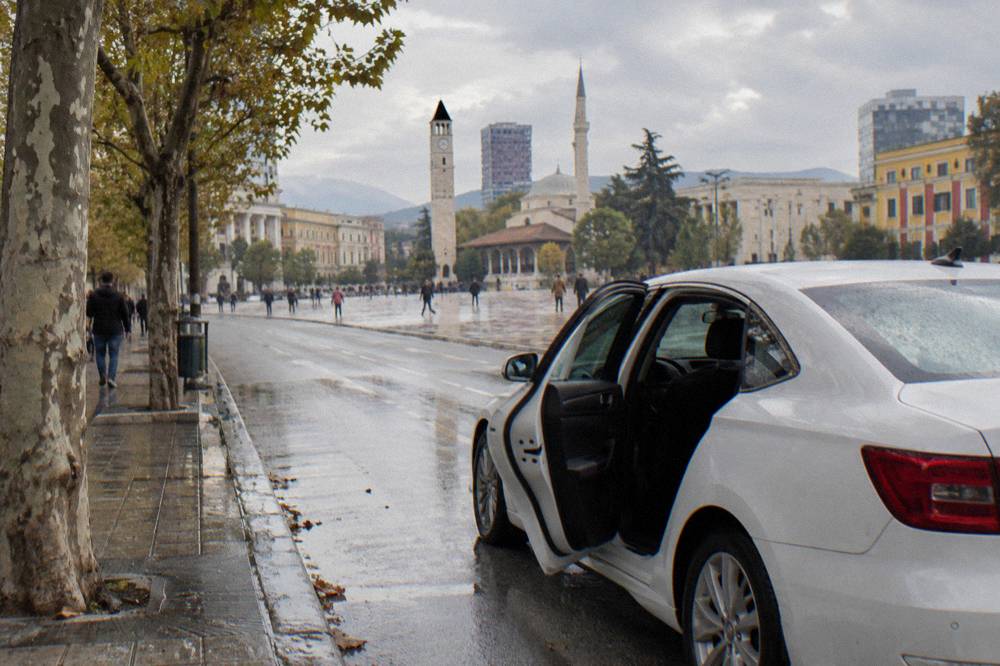 A white taxi waiting at a kerb on a tree-lined street in Tirana with its door open