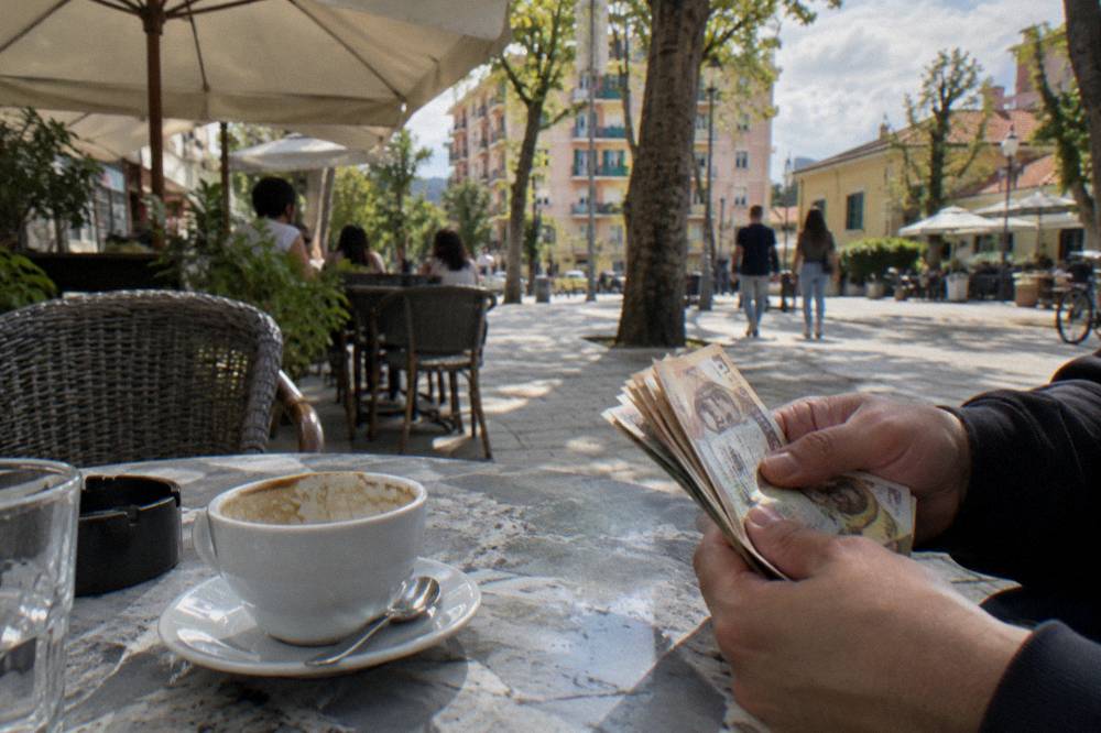 A hand counting Albanian lek banknotes across a cafe table beside a coffee cup