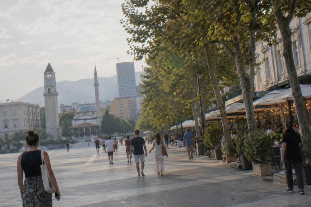 People strolling along a tree-lined Tirana boulevard at dusk with lit terraces behind