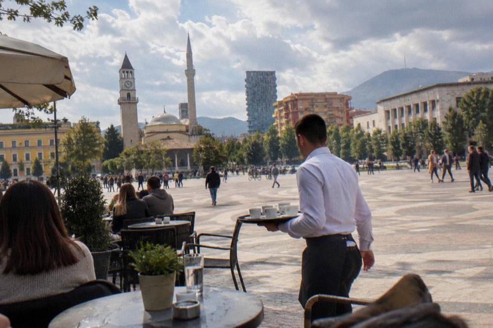 A waiter carrying a tray of espresso cups between tables on a busy Tirana terrace