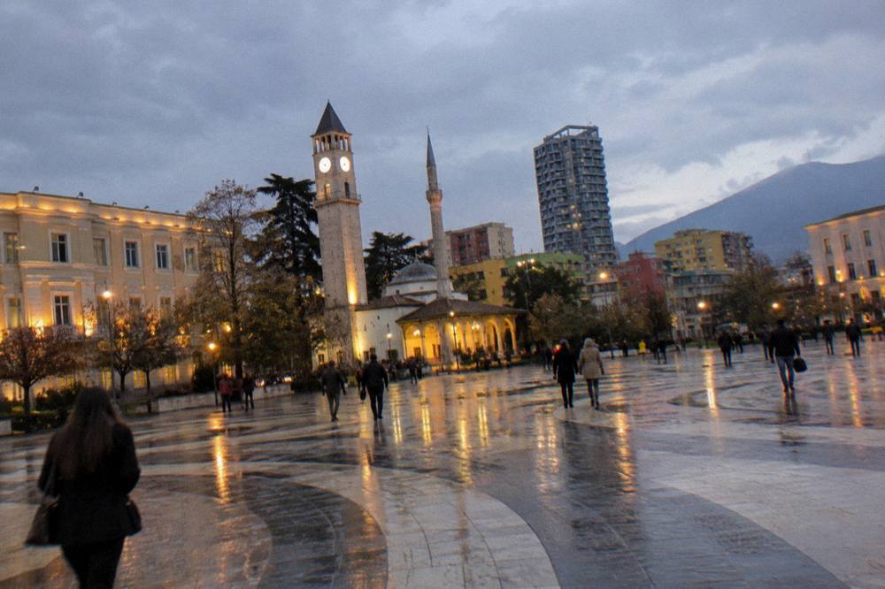 A city square at dusk with illuminated buildings and people walking