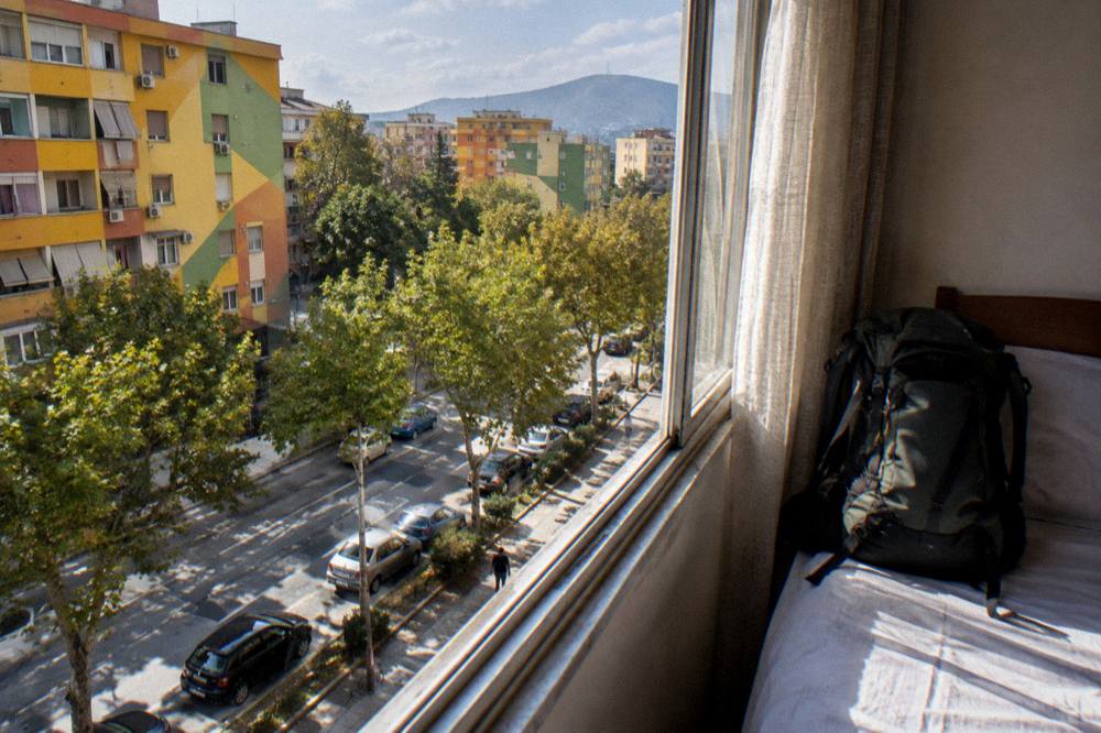 A packed backpack leaning against a hostel bed by a sunny window
