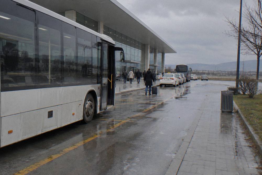 An airport shuttle bus parked at Tirana International Airport arrivals on an overcast morning
