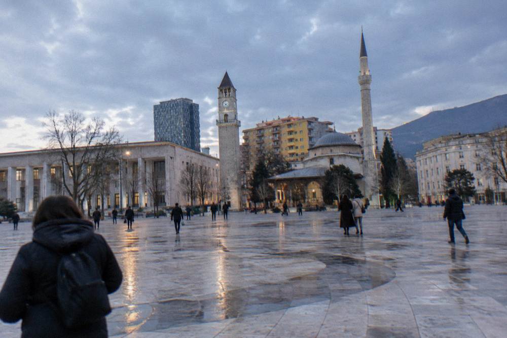 A wide pale square under a low grey sky with wet paving, bare trees and a slender stone tower