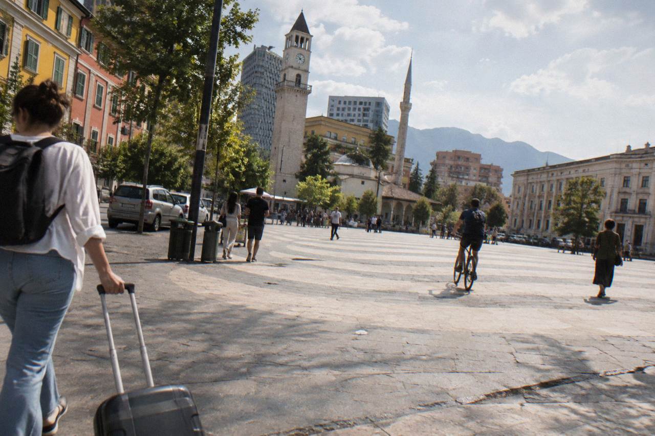 A traveller with a rolling suitcase walking along a sunny street of coloured buildings in central Tirana