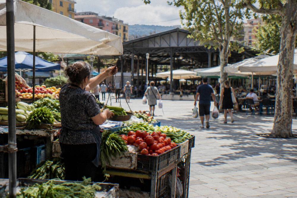 A vendor weighing vegetables on a hand scale at an outdoor market stall