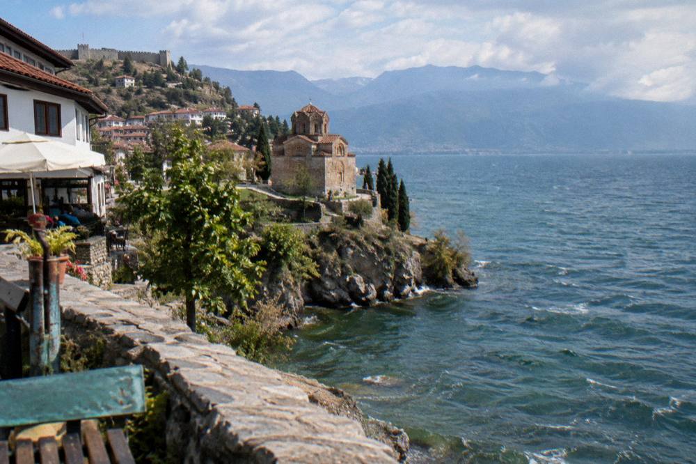 A small stone church on a cliff above a wide blue lake with mountains beyond