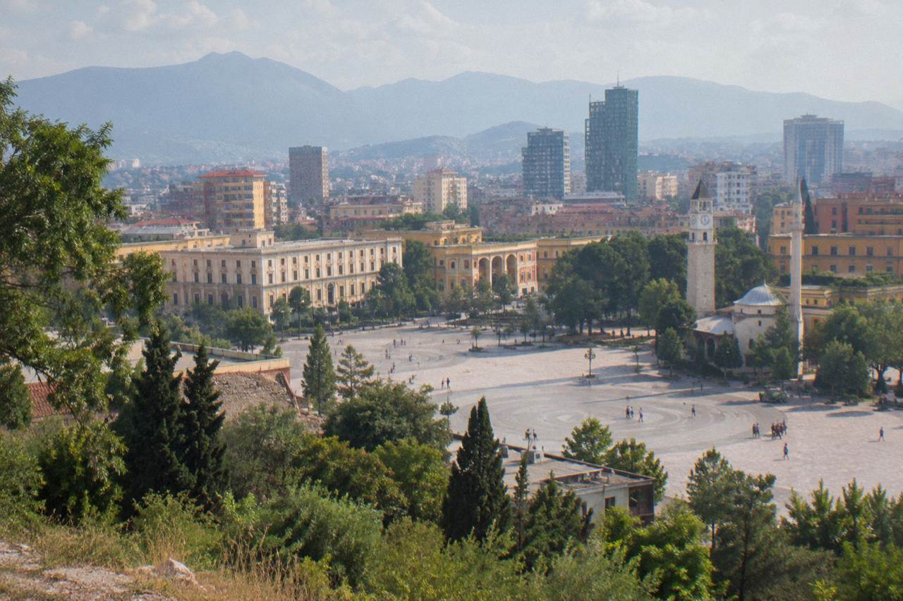 The city of Tirana seen from a high green slope, with rooftops and mountains behind