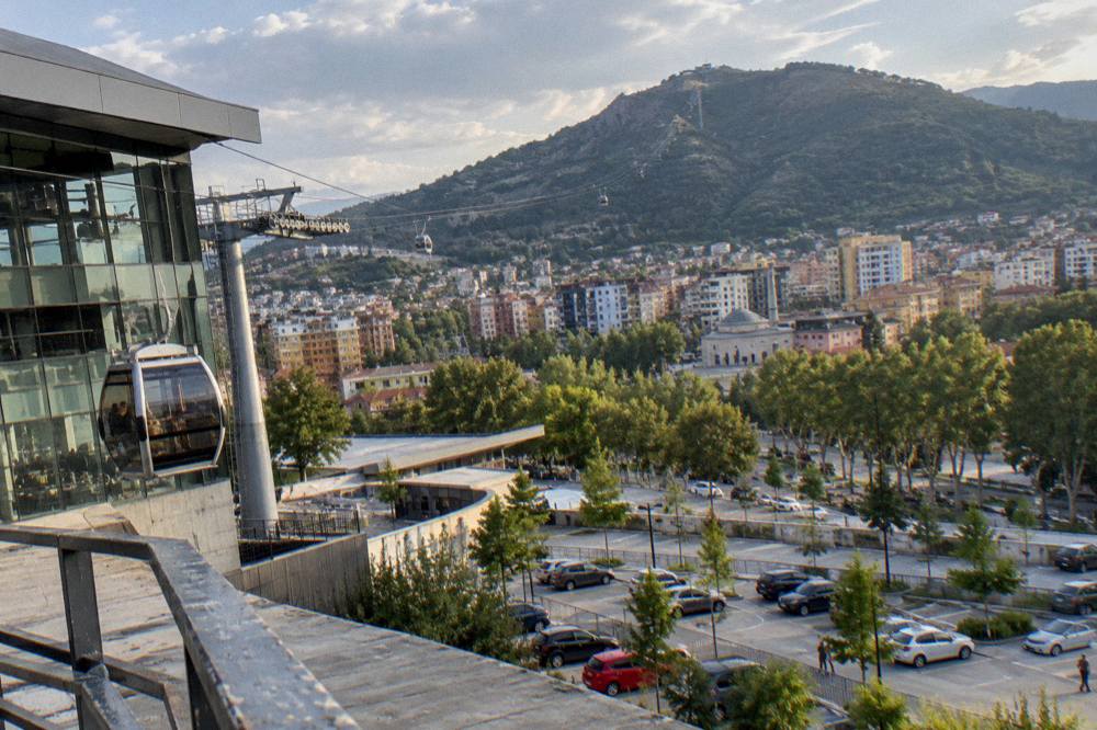 Evening view over city rooftops with a cable car line visible on a distant mountain