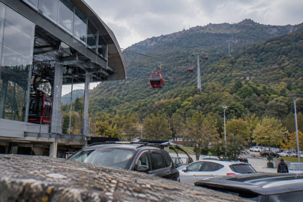 A red cable car cabin ascending above green slopes toward a mountain ridge above Tirana